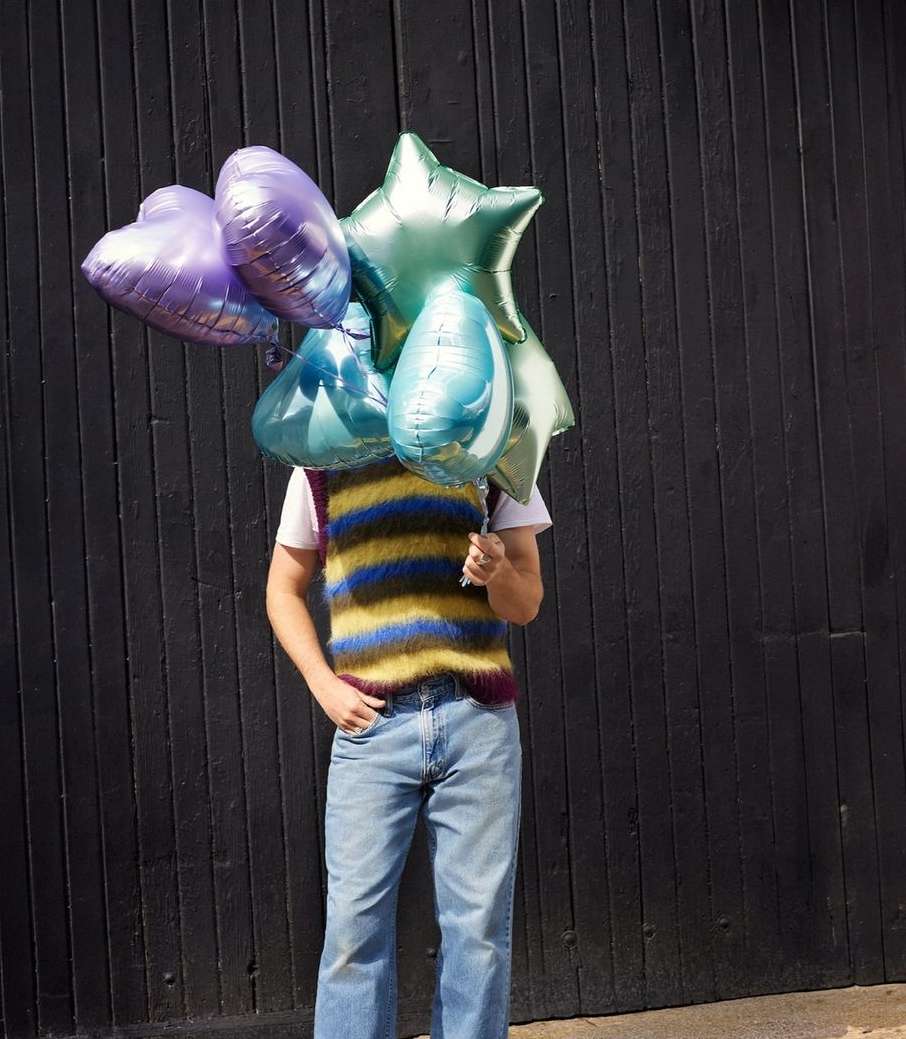 Person stands against a dark wood wall, their face hidden by a vibrant assortment of balloons in heart and star shapes.