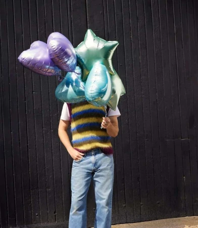 Person stands against a dark wood wall, their face hidden by a vibrant assortment of balloons in heart and star shapes.
