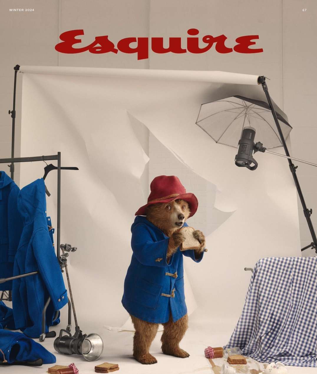 Paddington Bear in blue coat and red hat holds a sandwich in a photo studio with lighting equipment and prop table with checkered cloth.