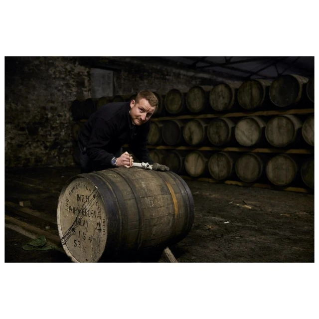 Man crouches near an aged oak barrel in a dimly lit, stone-walled cellar, showcasing traditional whiskey-making with rustic charm and craft.
