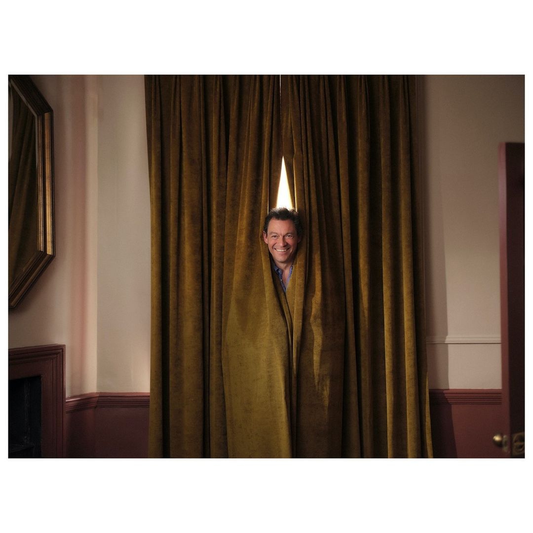 Smiling adult male peeks through gold velvet curtains indoors, with a dark wood fireplace and white walls visible in the background.
