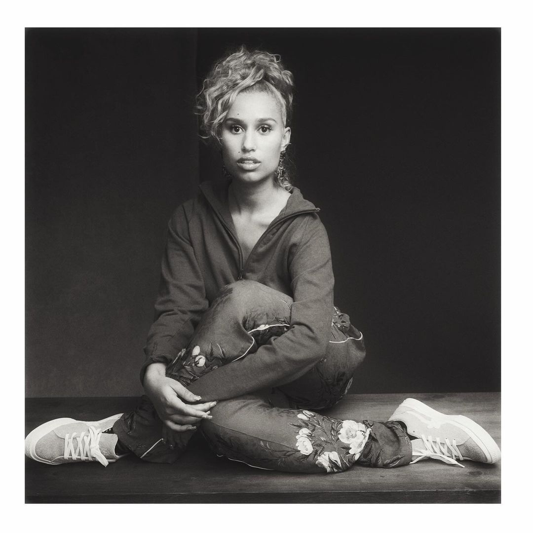 Portrait of a young adult female sitting cross-legged, wearing a hoodie and floral pants with white sneakers in a photo studio setting.