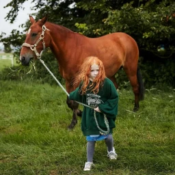 Young child with red hair wearing a dark green sweatshirt holding a lead rope attached to a brown horse standing in a grassy field