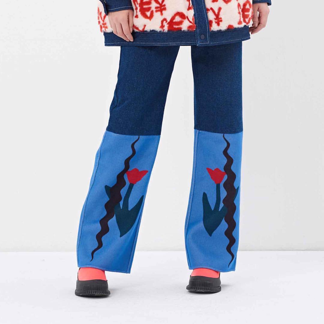Jeans with floral design worn by a person in a white background, complemented by dark shoes and a patterned cardigan.