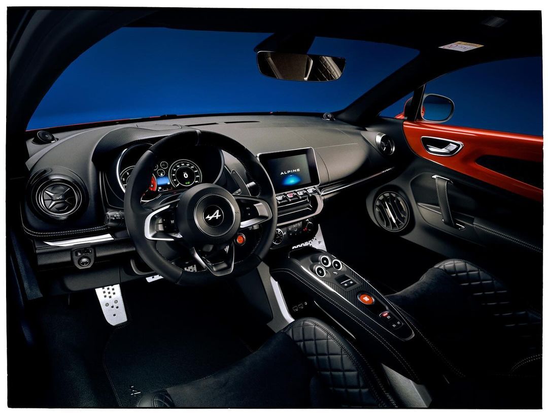 Interior of an orange Alpine sports car featuring a dashboard, steering wheel, black leather seats, and a sleek, modern design.