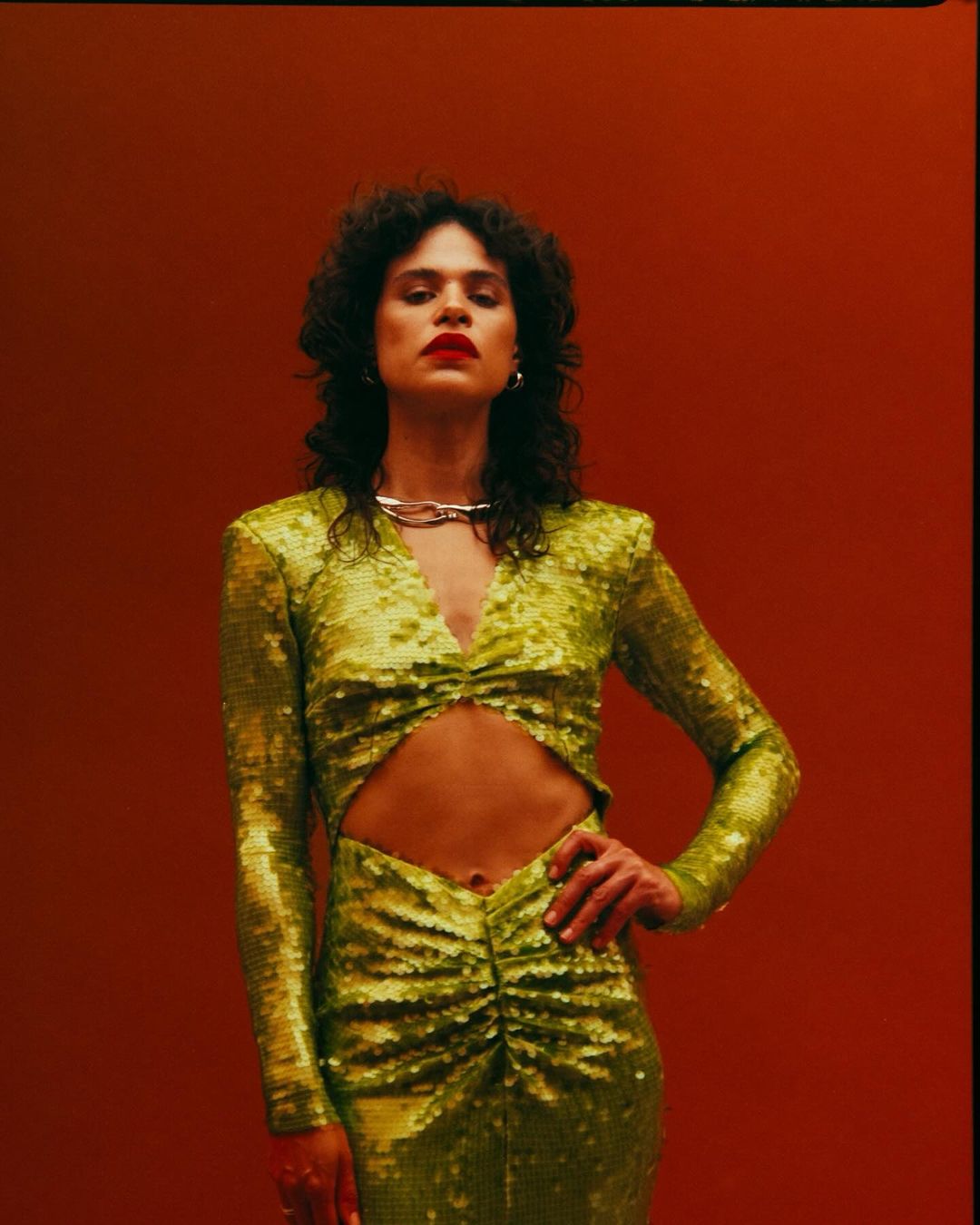 Woman in a sparkling green dress poses against a red backdrop, showcasing fashion and style, highlighting her dark hair and red lipstick.