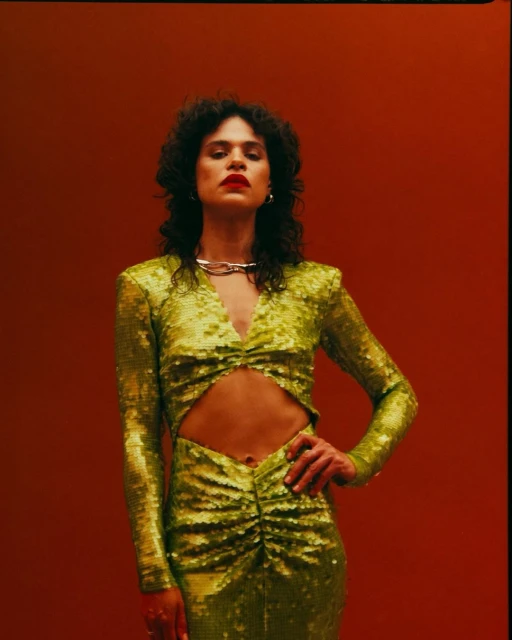 Woman in a sparkling green dress poses against a red backdrop, showcasing fashion and style, highlighting her dark hair and red lipstick.