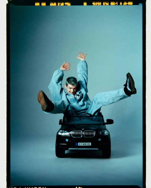 Man in denim outfit playfully poses in a child's black toy BMW X5 in a studio, arms raised and legs splayed against a gradient blue backdrop.
