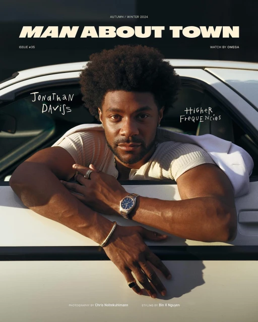 Man on the cover of 'Man About Town' magazine, leaning out of a car window, wearing a watch, bracelets, and rings, with natural lighting.
