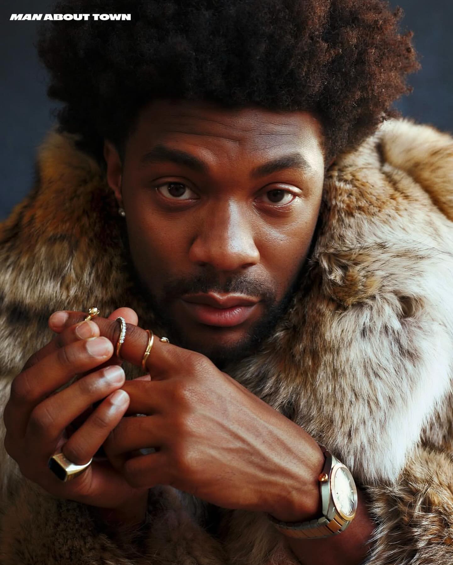 Portrait of a Black man with an afro wearing a fur-trim coat, rings, and wristwatch, photographed by Chris Noltekuhlmann.