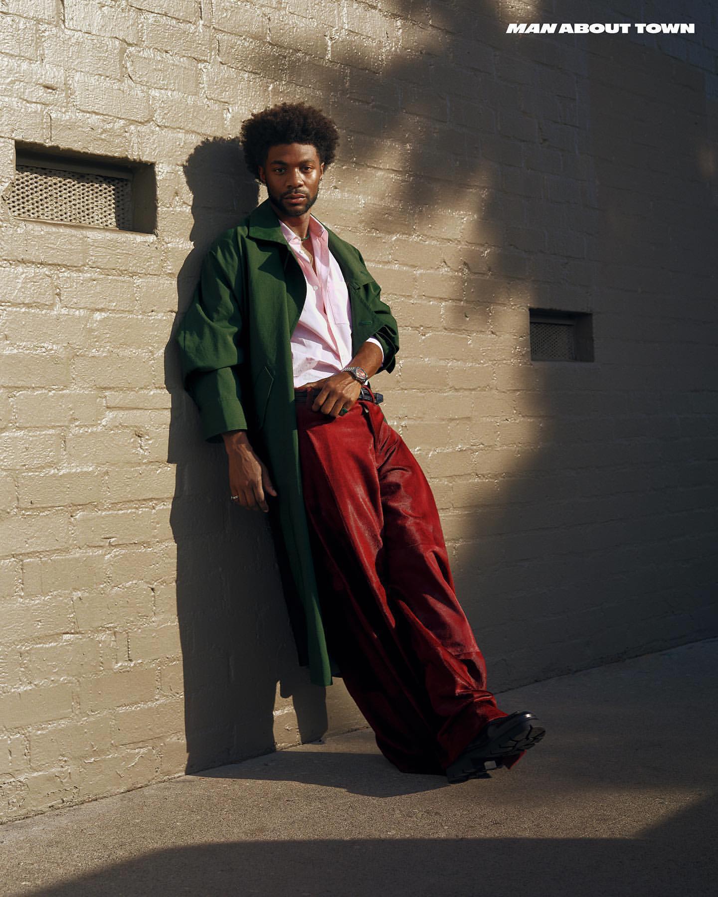 A stylish, curly-haired man in a green coat and red pants leans against a textured wall in dappled sunlight, exuding urban fashion.