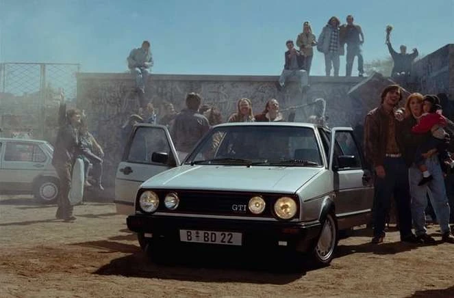 Silver Volkswagen Golf GTI parked in front of a crowd of people and a graffiti-covered wall under a clear sky on a sunny day.