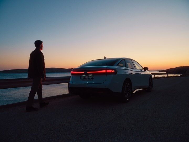 A man walks along a coastal road at sunset, with a modern white sedan parked nearby, silhouetted against the colorful sky and ocean.