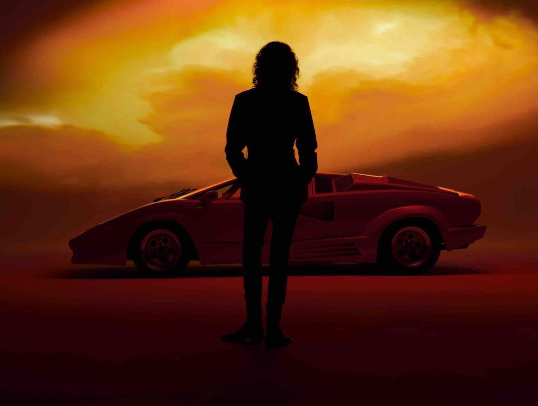 Silhouette of a man standing in front of a red Lamborghini against a gradient orange and yellow sky, creating a striking contrast.
