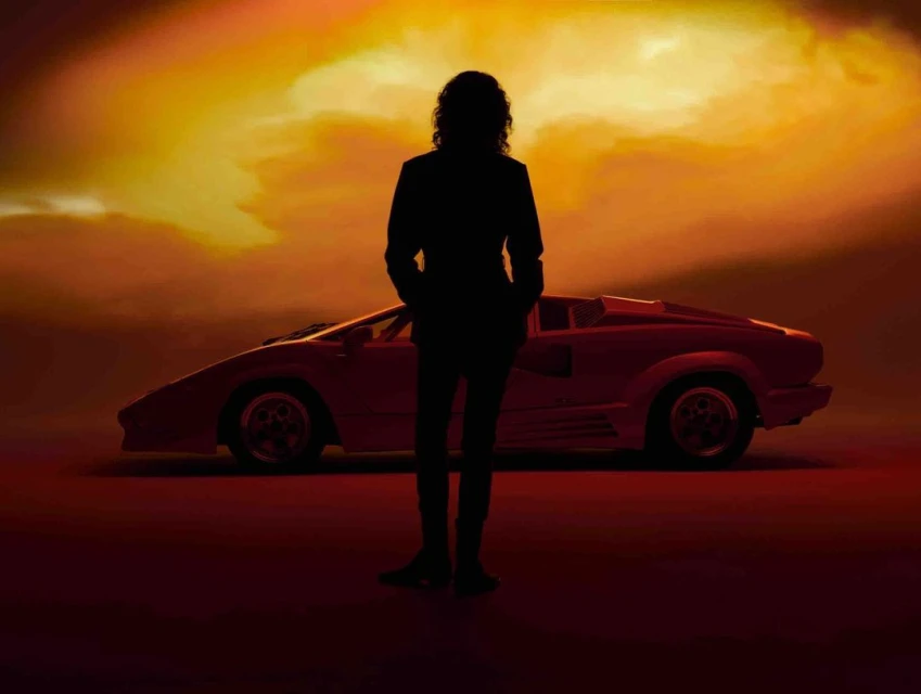 Silhouette of a man standing in front of a red Lamborghini against a gradient orange and yellow sky, creating a striking contrast.