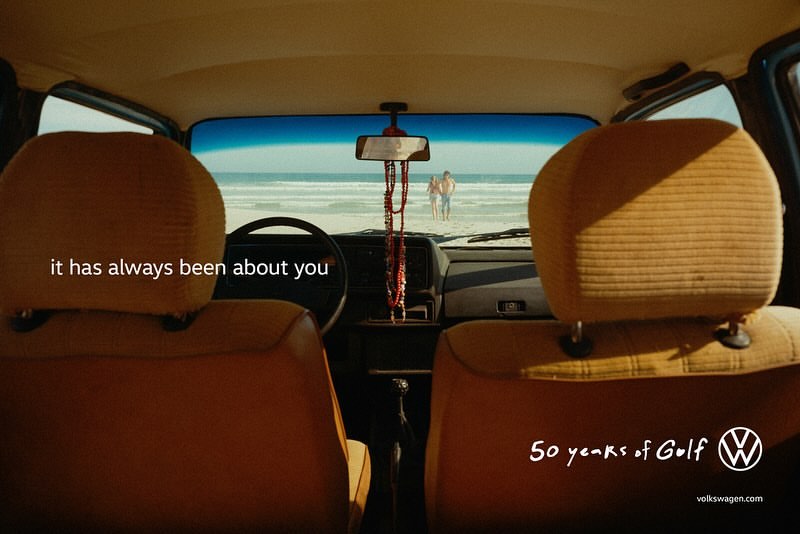 Car interior view looking out at a beach with two people, calm ocean water, and clear sky, Volkswagen advertisement for the 50th year.