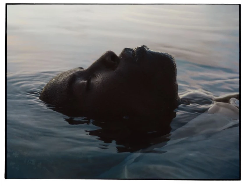 Close-up of a person's dark-skinned face, submerged in calm water, with soft natural light casting a gentle glow on the scene.