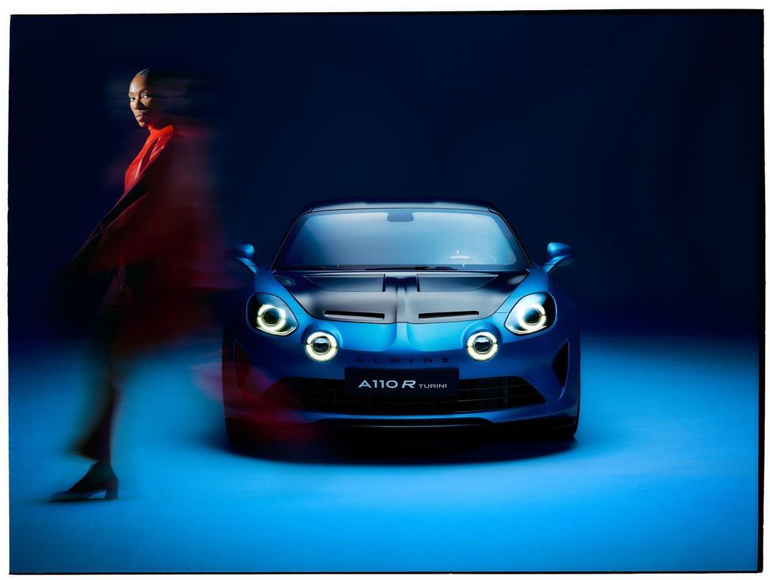 A blue Alpine A110 R Turini car is parked in a studio with a blurred woman walking by in the background creating motion.