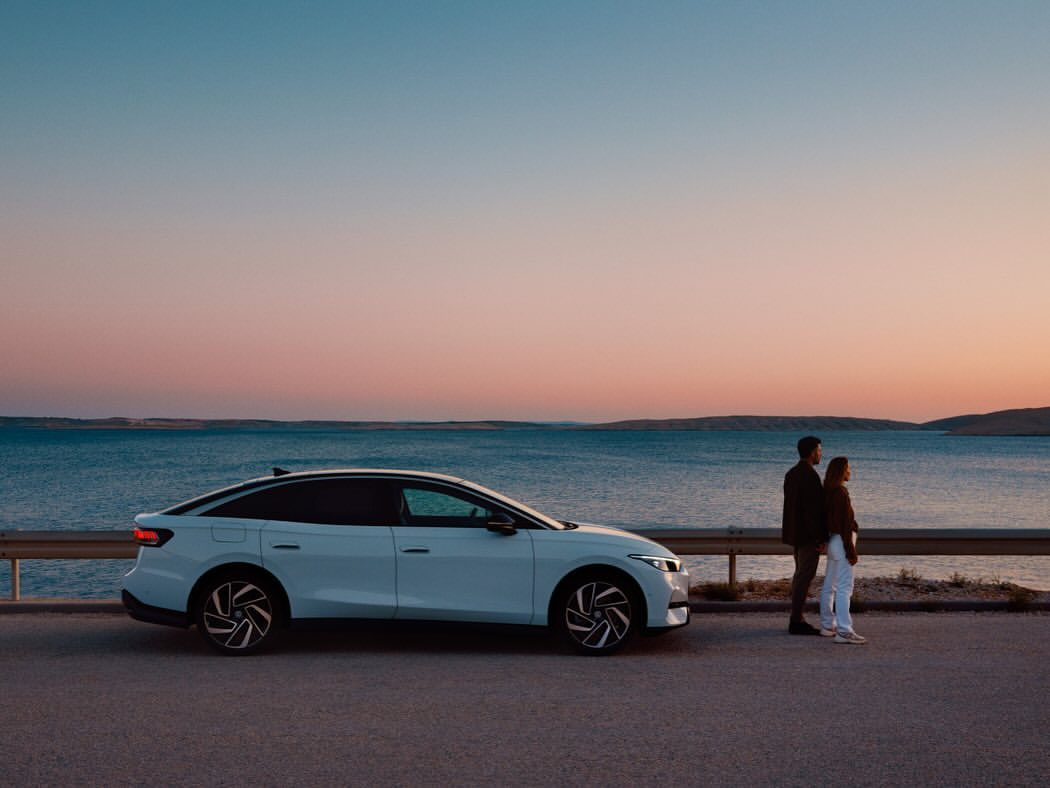 White Volkswagen ID.7 electric vehicle parked near a couple standing by the sea at sunset, offering a blend of automotive and lifestyle.