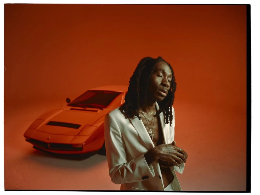 Stylish adult male in a white suit poses in front of a classic white sportscar against a solid orange backdrop in a studio setting.