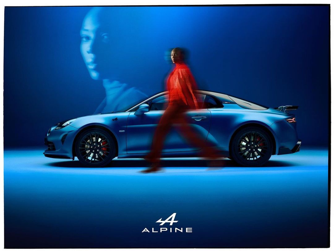 A blue Alpine sports car is parked indoors while a blurry figure walks past it, with a woman's face projected in the background.