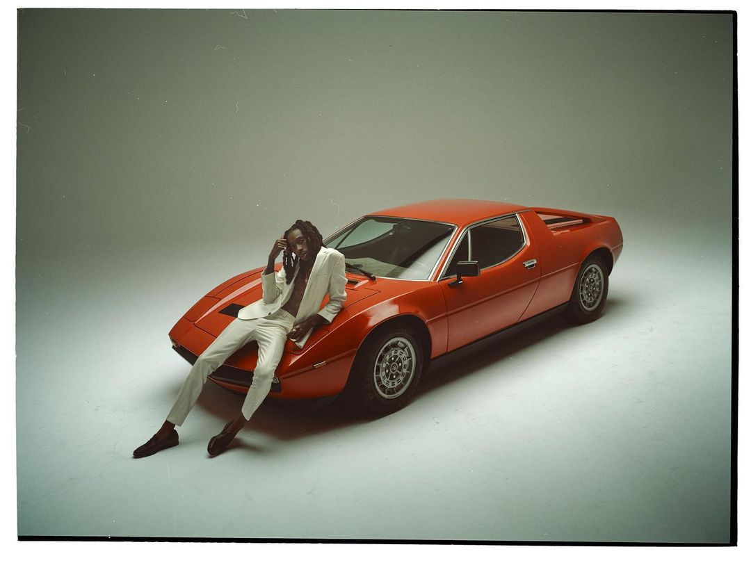 Man in a white suit poses against a bright orange Maserati car in a studio setting, showcasing fashion and automotive design.