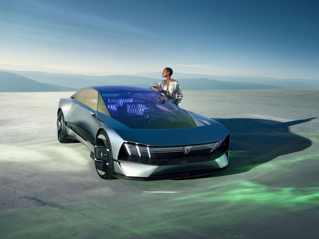 Sleek silver car on expansive concrete surface with woman leaning casually against it, distant mountains under blue sky.