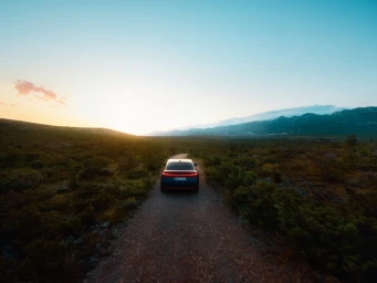 Rear view of a modern electric car driving down a narrow dirt road through an open landscape under a dramatic sunset sky.