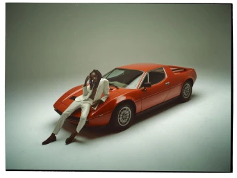 Man in a white suit leaning against a vintage red sports car in a studio setting with grey seamless background