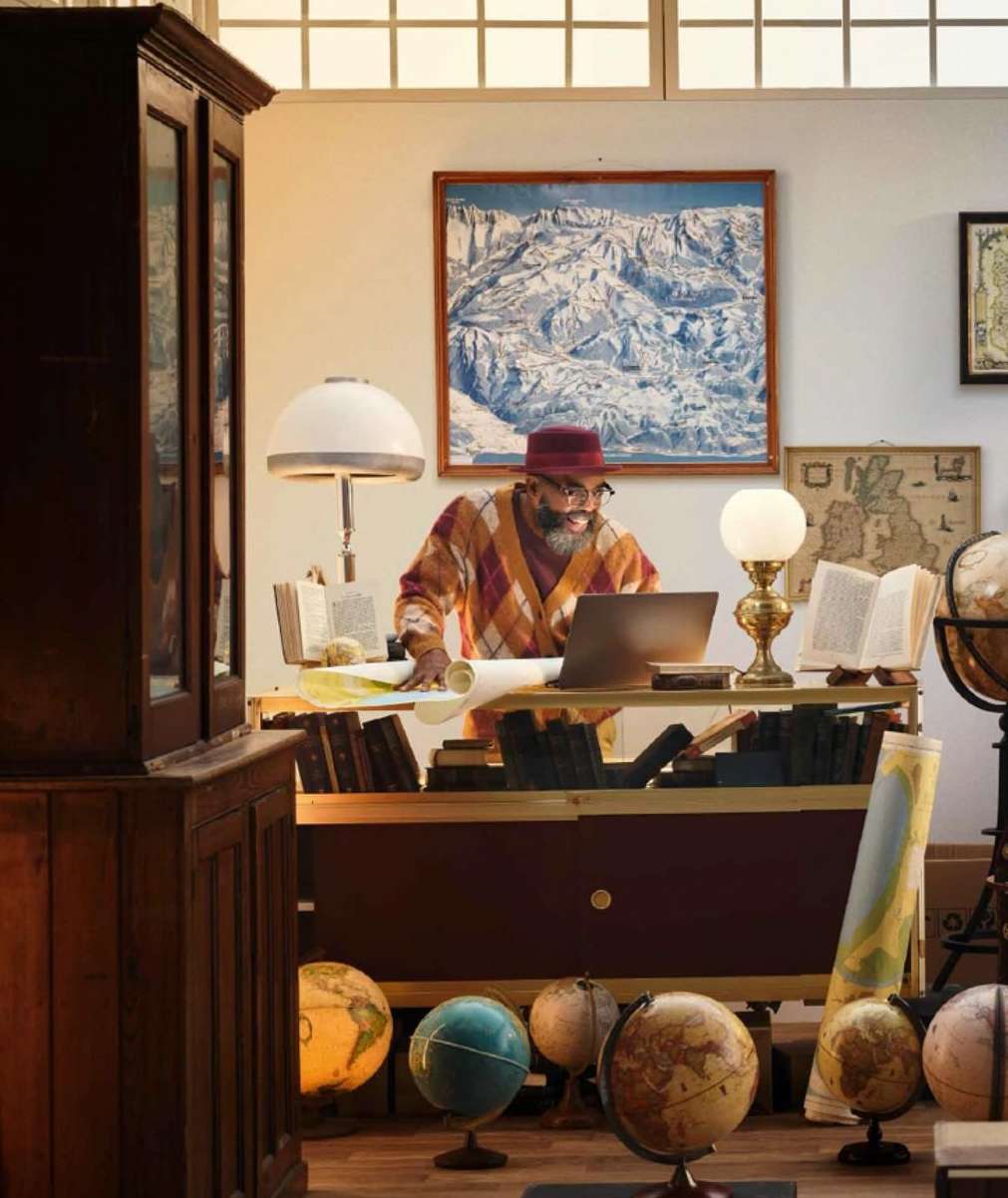 Adult man wearing glasses, hat, and orange sweater working on a laptop on a desk with maps and globes in a room with books.