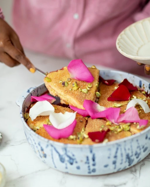 Woman in pink blouse holding a plate and using a spatula to serve semolina cake decorated with rose petals and pistachios.