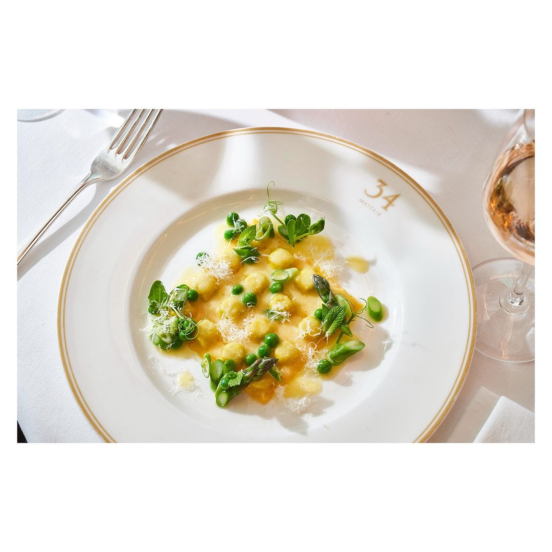 Gourmet dish featuring pasta, peas, and asparagus on a white plate with gold trim, alongside a fork and a glass of rose wine.