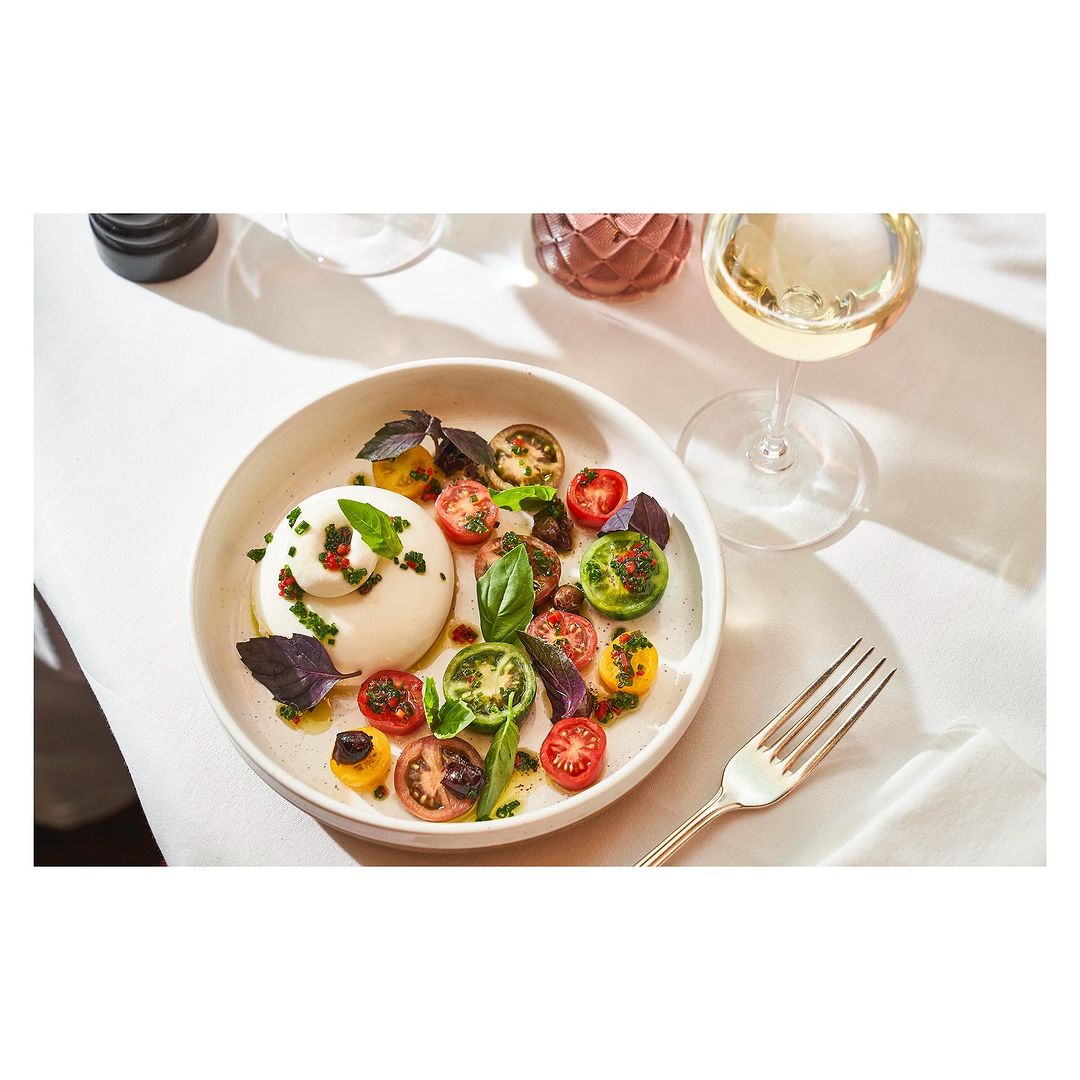 Overhead shot of burrata salad with cherry tomatoes, basil, and balsamic glaze on a white plate, next to a glass of white wine.