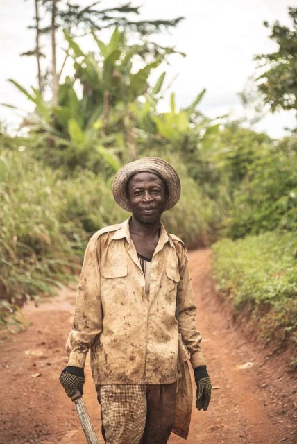 African man in straw hat and work clothes stands on a dirt road surrounded by green vegetation. Outdoor portrait on a cloudy day.
