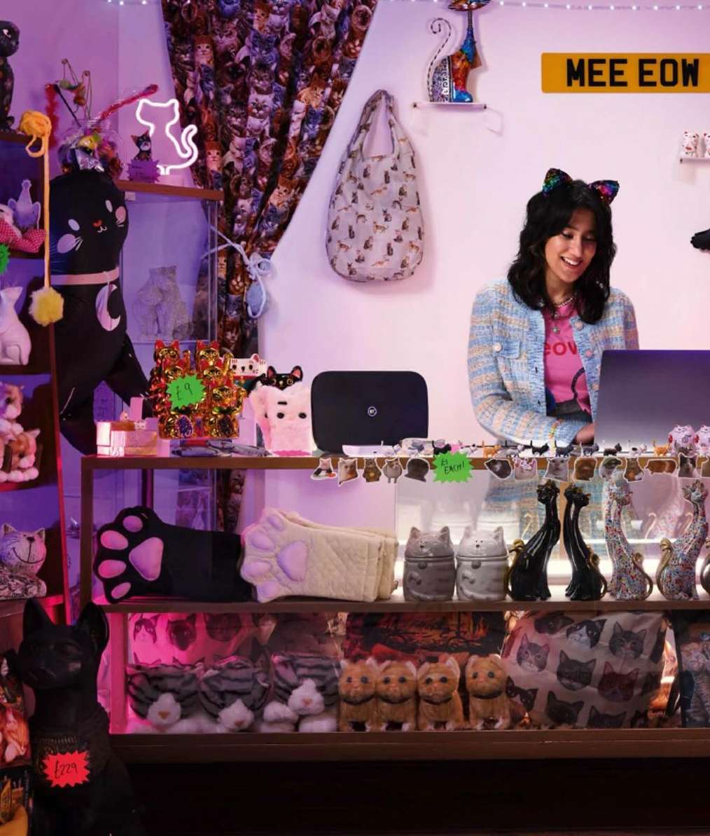 Woman with cat-ear headband stands behind counter laden with cat-themed objects, illuminated by pink lighting, in shop.