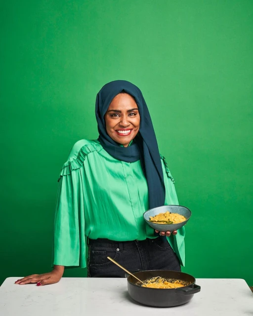 Adult female wearing a hijab and a green top holding a bowl of yellow food, next to a pot of food on a white table with a green background.