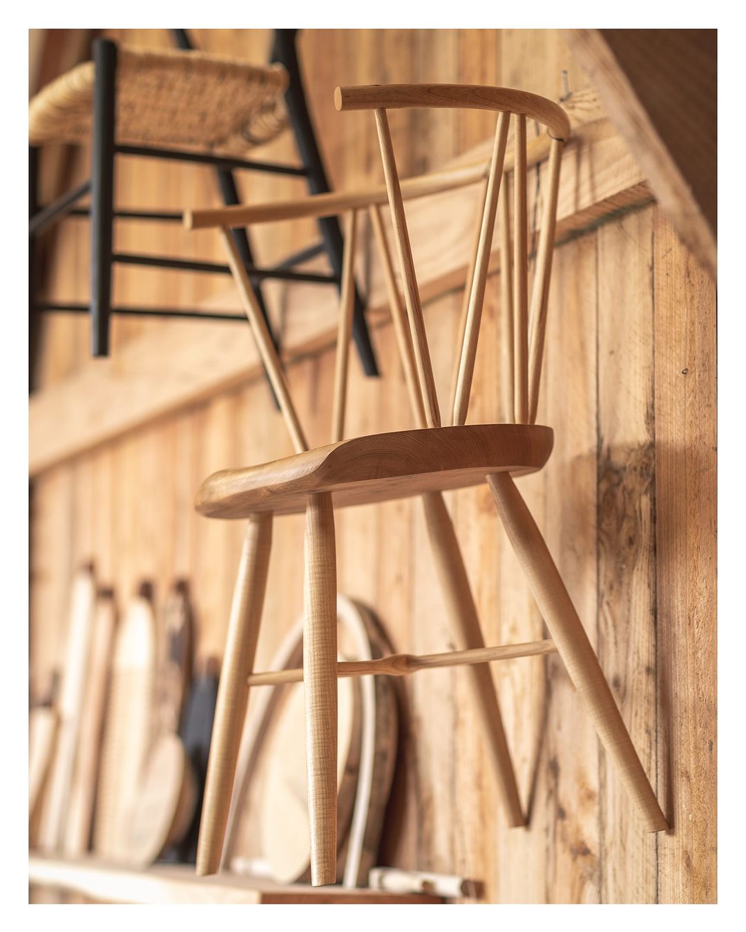 Close-up of a light wood chair, crafted with slender spindles, positioned against a wall with additional woodworking pieces.