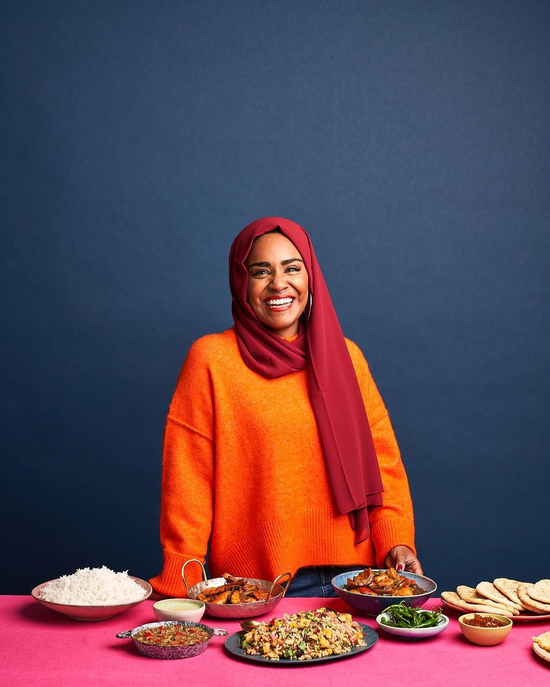 Smiling woman in orange sweater and burgundy hijab stands behind table of assorted prepared foods against dark blue studio backdrop.