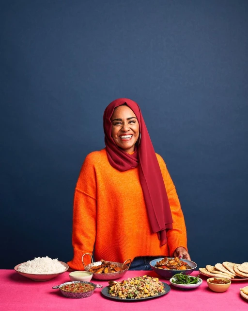 Smiling woman in orange sweater and burgundy hijab stands behind table of assorted prepared foods against dark blue studio backdrop.