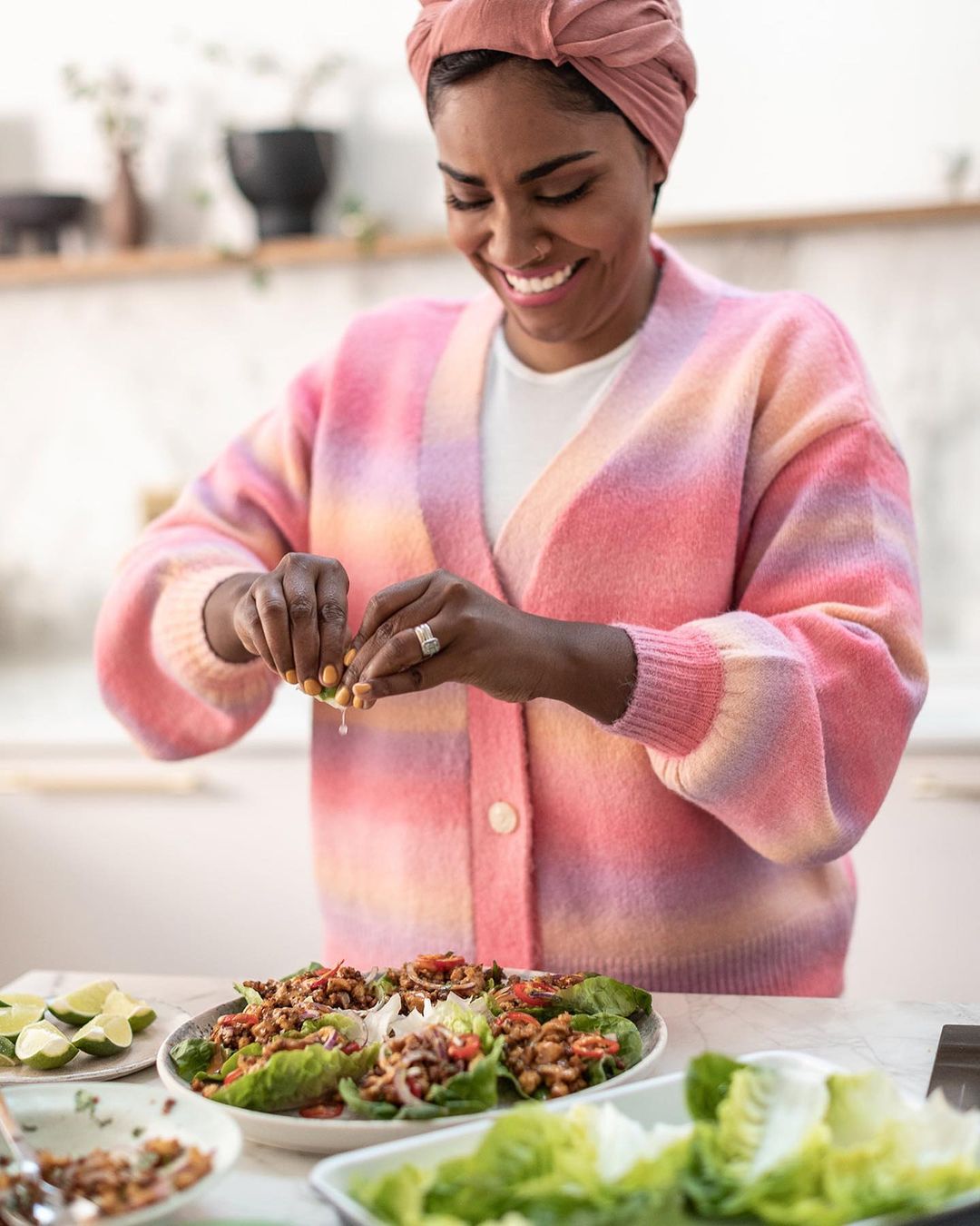 Woman squeezes lime juice onto food on lettuce leaves in bright kitchen. She wears a pink headscarf and pink tie-dye sweater.