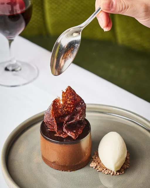 Elegant dessert presentation: chocolate mousse, ice cream, and garnish on a grey plate, being drizzled with liquid from a spoon.