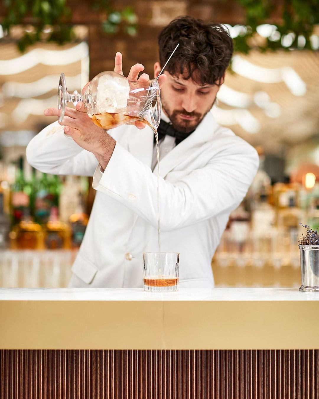 A bartender wearing a white jacket mixes a cocktail at a bar. The bartender strains a drink into a glass with ice, indoors, bar setting.
