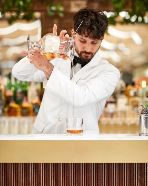 A bartender wearing a white jacket mixes a cocktail at a bar. The bartender strains a drink into a glass with ice, indoors, bar setting.
