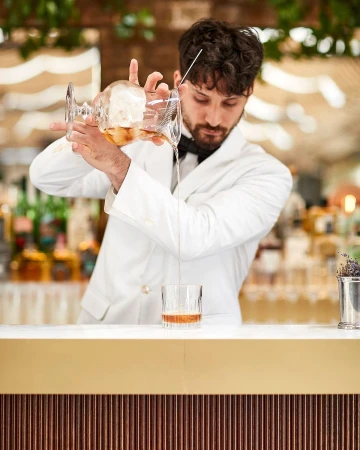 A bartender wearing a white jacket mixes a cocktail at a bar. The bartender strains a drink into a glass with ice, indoors, bar setting.