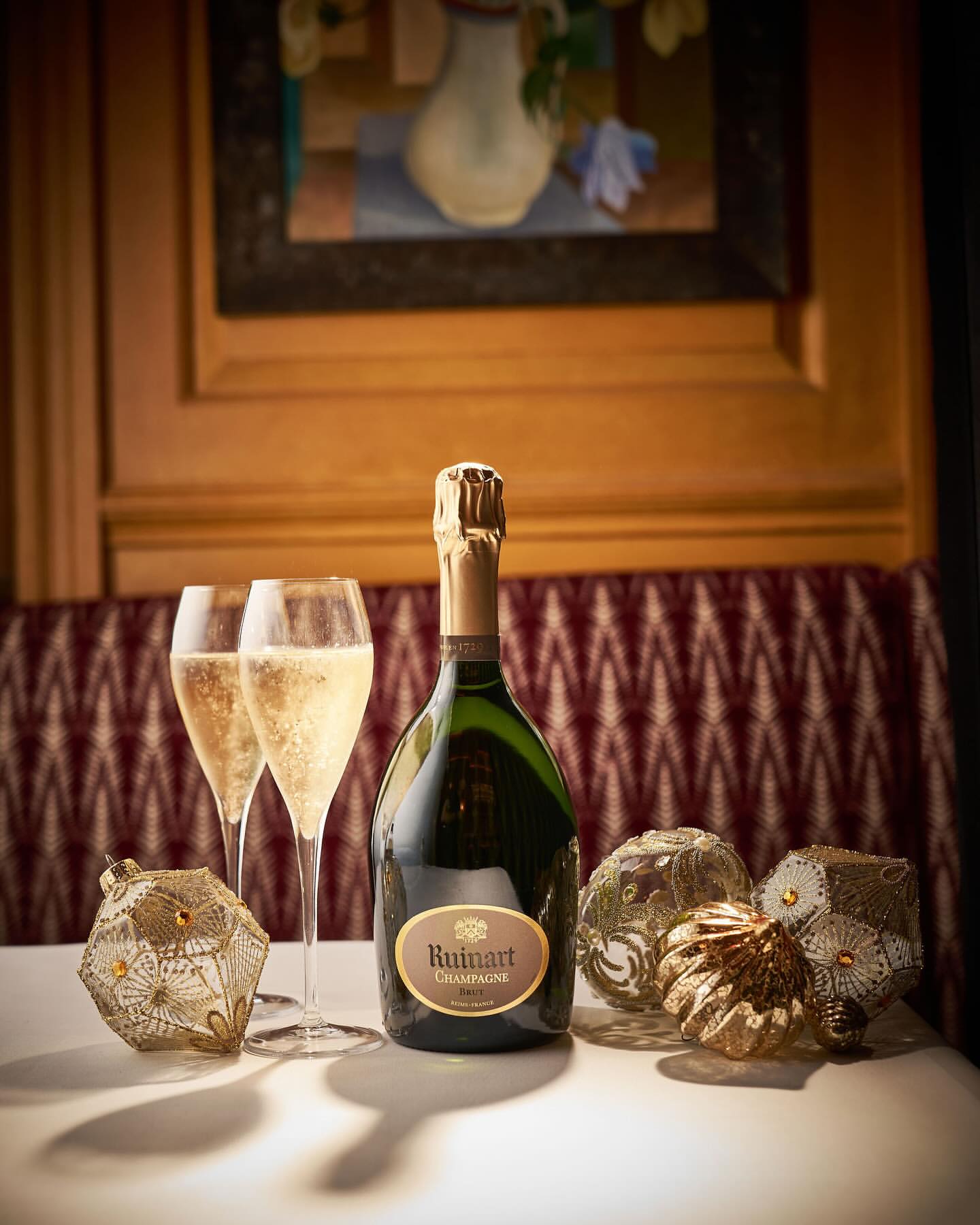 Bottle of Ruinart Champagne with two glasses of sparkling wine and decorative ornaments on a white table with a painting in background.
