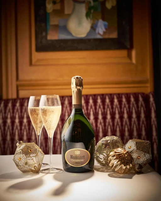 Bottle of Ruinart Champagne with two glasses of sparkling wine and decorative ornaments on a white table with a painting in background.