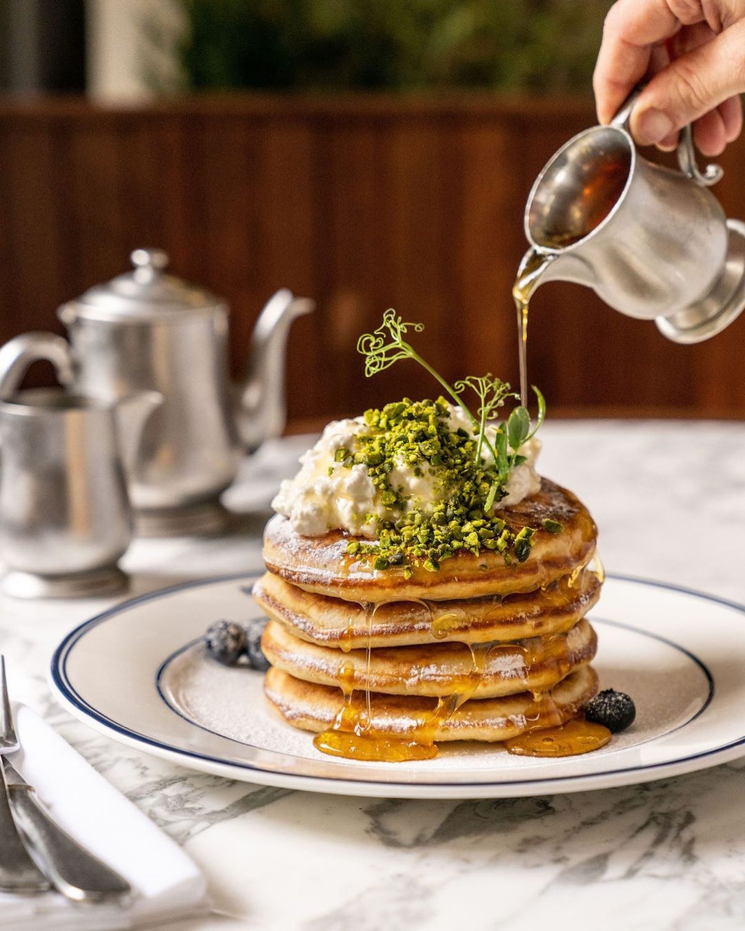 Stack of pancakes topped with cream and pistachios being drizzled with syrup next to silverware and a silver teapot.