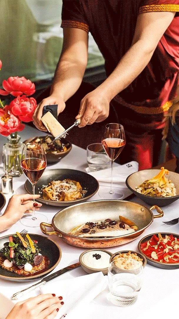 Overhead shot of a catered table with a chef grating cheese over a dish surrounded by rose wine and multiple plates of food.