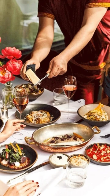 Overhead shot of a catered table with a chef grating cheese over a dish surrounded by rose wine and multiple plates of food.