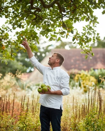 A chef in a white coat stands in a garden under a tree, reaching for apples. He is holding green apples in his arm.